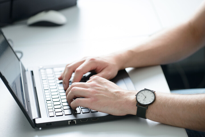 aufgeschlagener Laptop mit zwei Händen an der Tastatur, am rechten Handgelenk befindet sich eine Armbanduhr.
