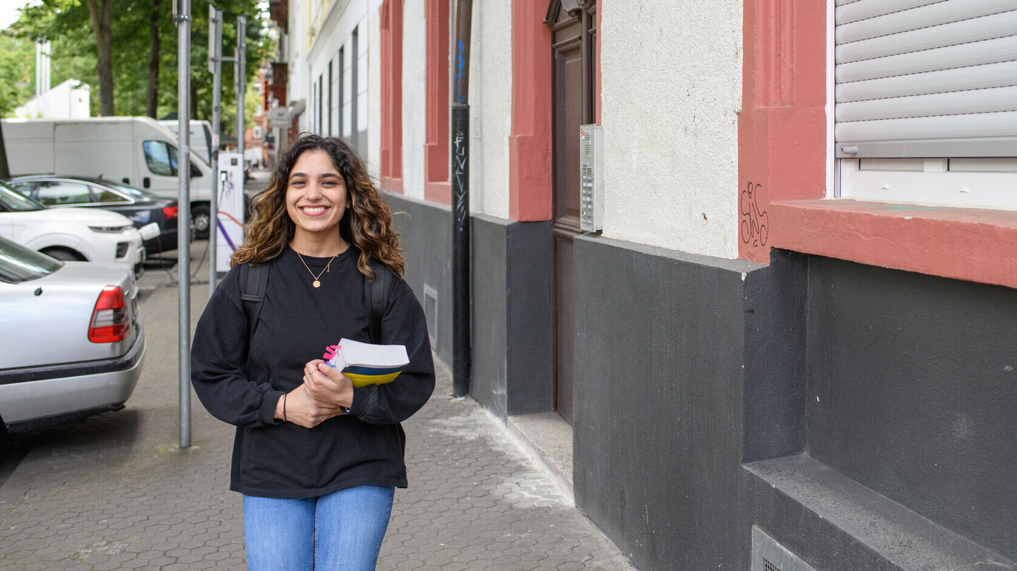 Ein*e Student*in läuft durch eine Straße, im Hintergrund eine Hausfassade und parkende Autos. Die Person trägt ein dickes Buch, hat lange gewellte Haare und lächelt.
