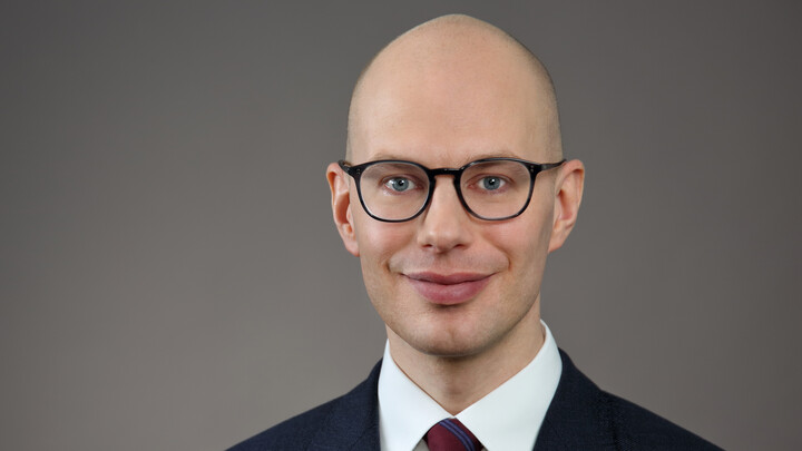Eine Person trägt ein weißes Hemd, einen dunkelblauen Blazer sowie eine rot blau gestreifte Krawatte und steht vor einem grauen Hintergrund. Die Person heißt Nils Schaks.