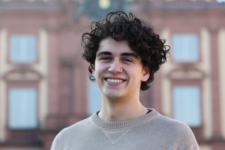 Sebastian has curly black hair and a nose piercing. He wears a beige pullover and stands in front of the Schloss.