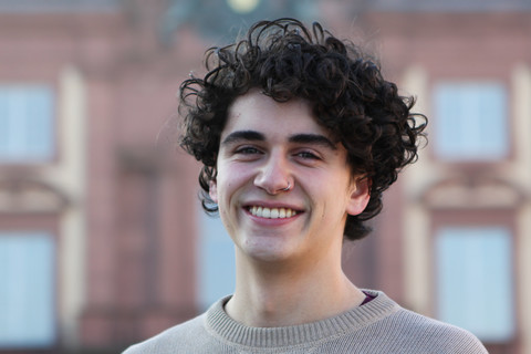 Sebastian has curly black hair and a nose piercing. He wears a beige pullover and stands in front of the Schloss.