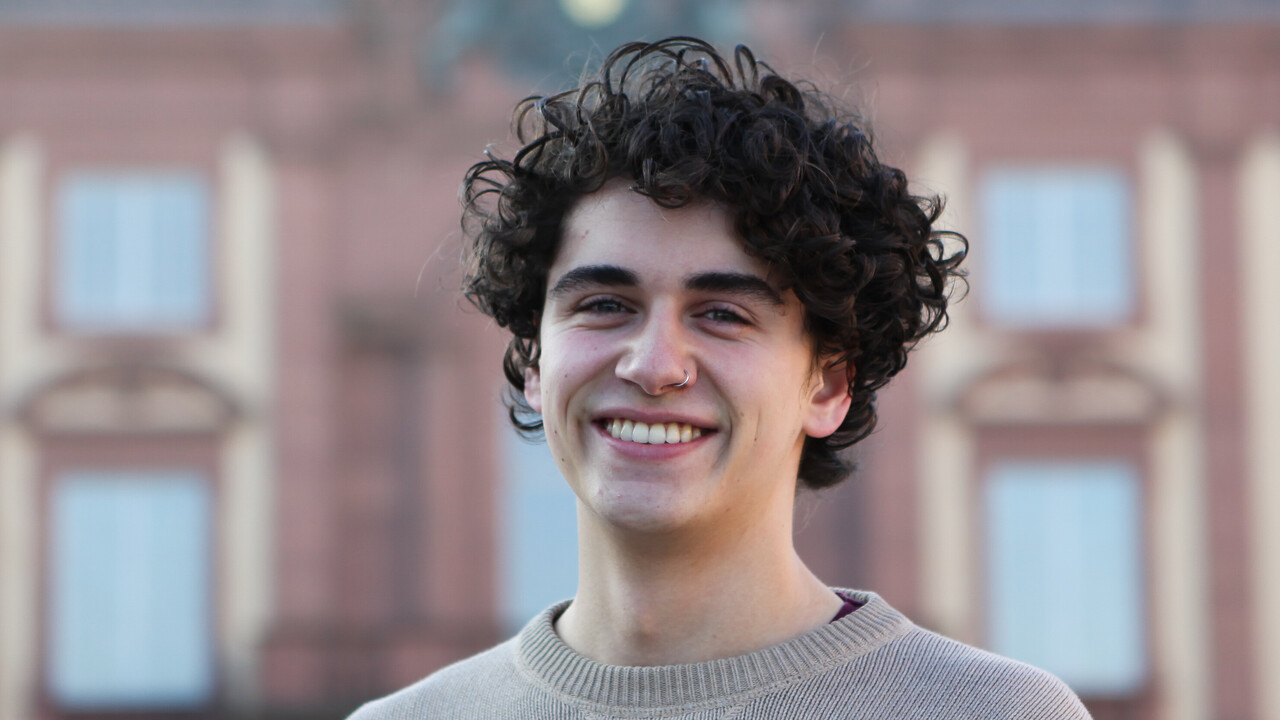 Sebastian has curly black hair and a nose piercing. He wears a beige pullover and stands in front of the Schloss.