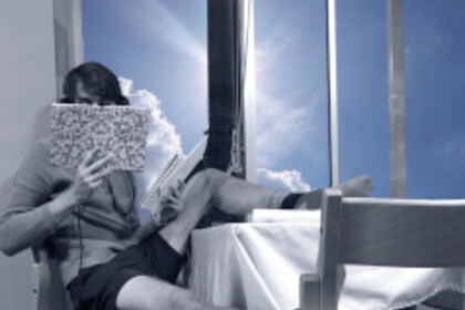 Ein Mann in Shorts und Pulli sitzt auf einem Stuhl am Fenster, einen Fuß auf dem Tisch, den anderen auf einem Stuhl, und liest in einem Buch.