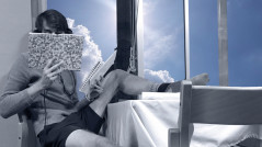 Ein Mann in Shorts und Pulli sitzt auf einem Stuhl am Fenster, einen Fuß auf dem Tisch, den anderen auf einem Stuhl, und liest in einem Buch.