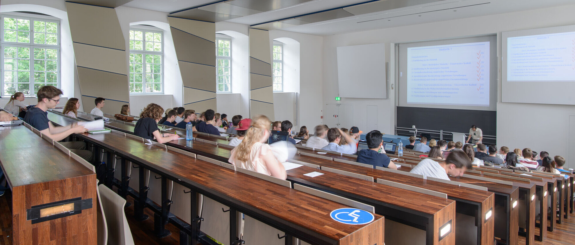 Großer Hörsaal mit vielen Studierenden. Vorne schaut ein Dozent in seine Unterlagen. 