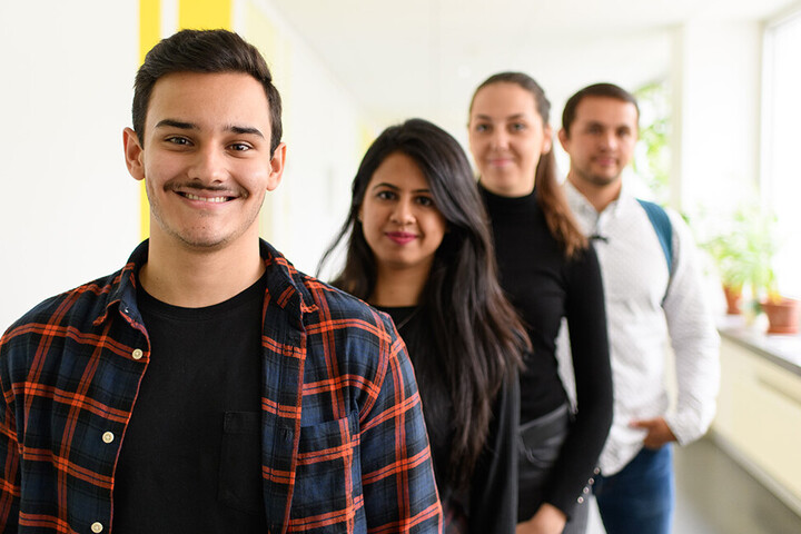 Vier Studierende stehen in einer Reihe versetzt hinterheinander und lachen in die Kamera.
