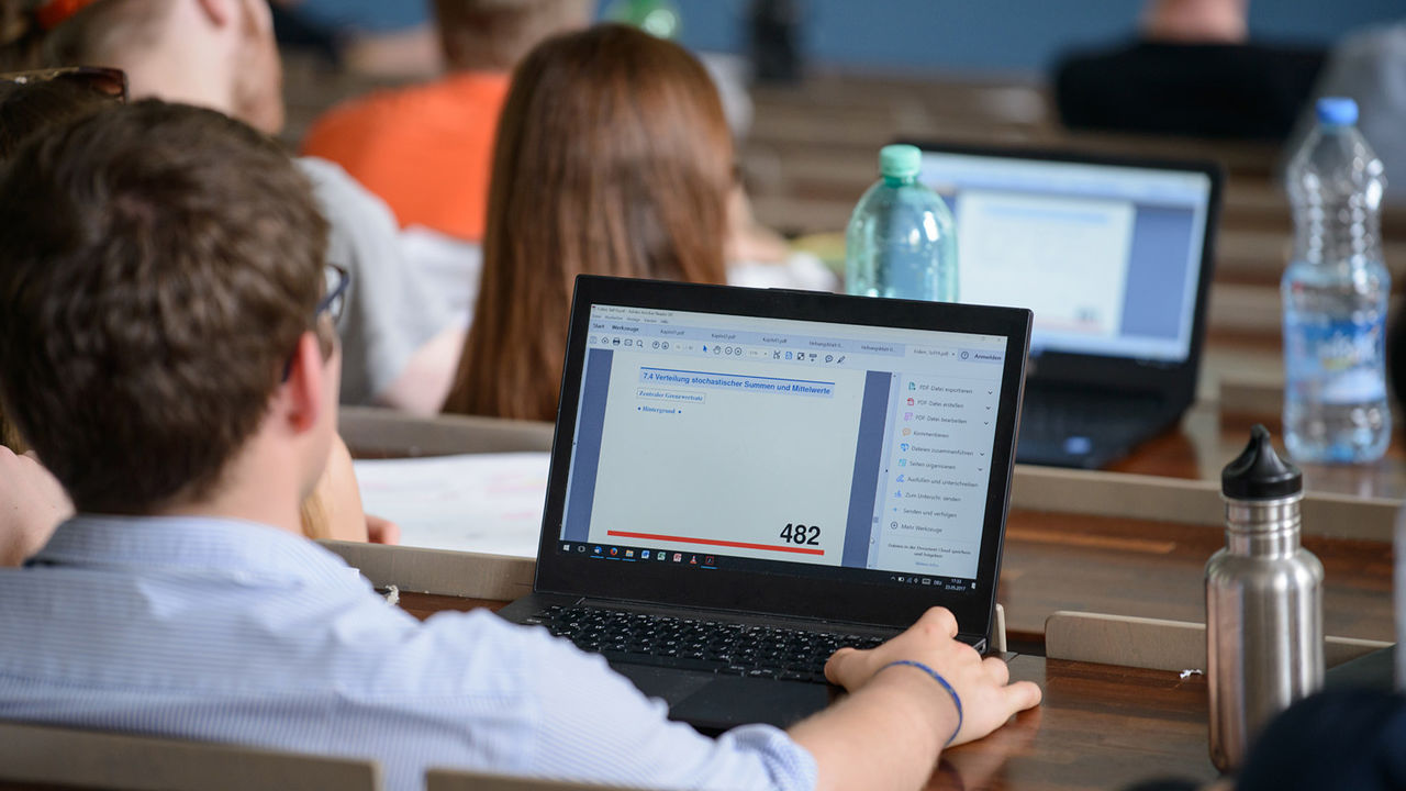 Ein Student sitzt in einem Hörsaal und tippt am Laptop. Er trägt eine braune Kurzhaarfrisur, eine Brille und ein blaues Hemd.