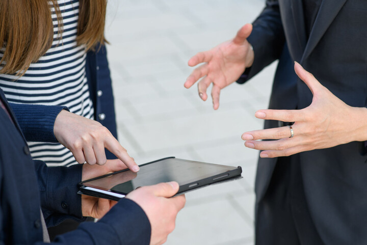 Eine Person hält ein Tablet in den Händen. Eine zweite Personen zeigt auf den schwarzen Bildschirm des Tablets. Eine dritte Persob gestikuliert mit den Händen in der Luft.