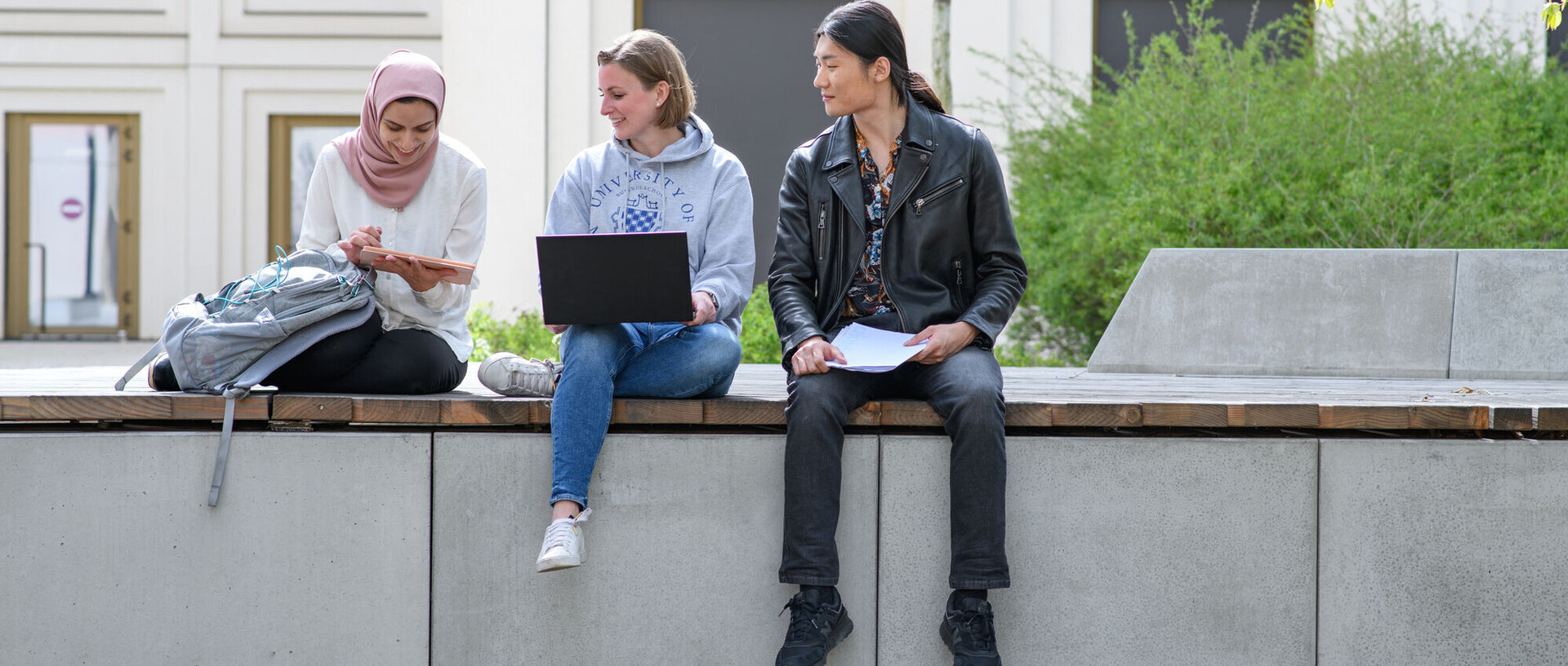 Drei Studierende sitzen draußen auf dem Unigelände mit Laptop, Tablet und Unterlagen und schauen sich etwas auf dem Tablet an.