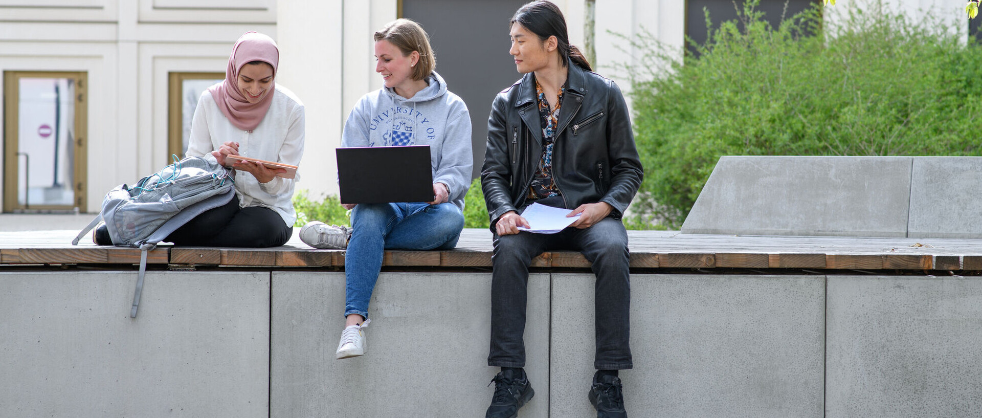 Drei Personen sitzen draußen auf dem Unigelände mit Laptop, Tablet und Unterlagen und schauen sich etwas auf dem Tablet an.