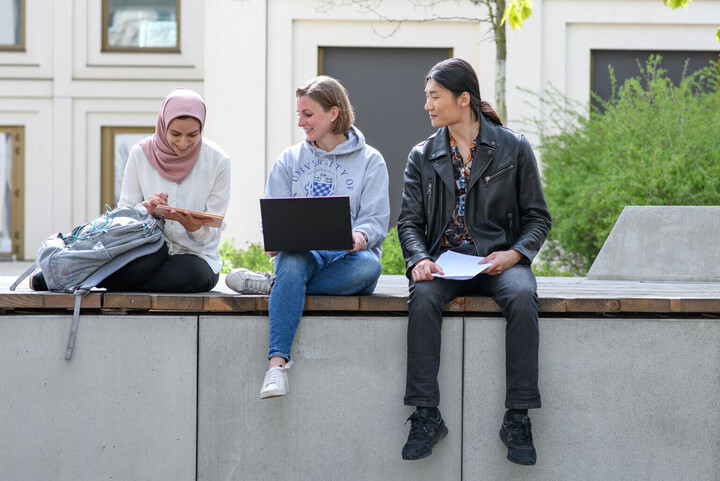 Drei Studierende sitzen draußen auf dem Unigelände mit Laptop, Tablet und Unterlagen und schauen sich etwas auf dem Tablet an.