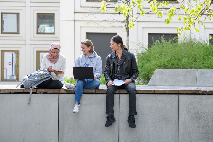 Drei Studierende sitzen draußen auf dem Unigelände mit Laptop, Tablet und Unterlagen und schauen sich etwas auf dem Tablet an.