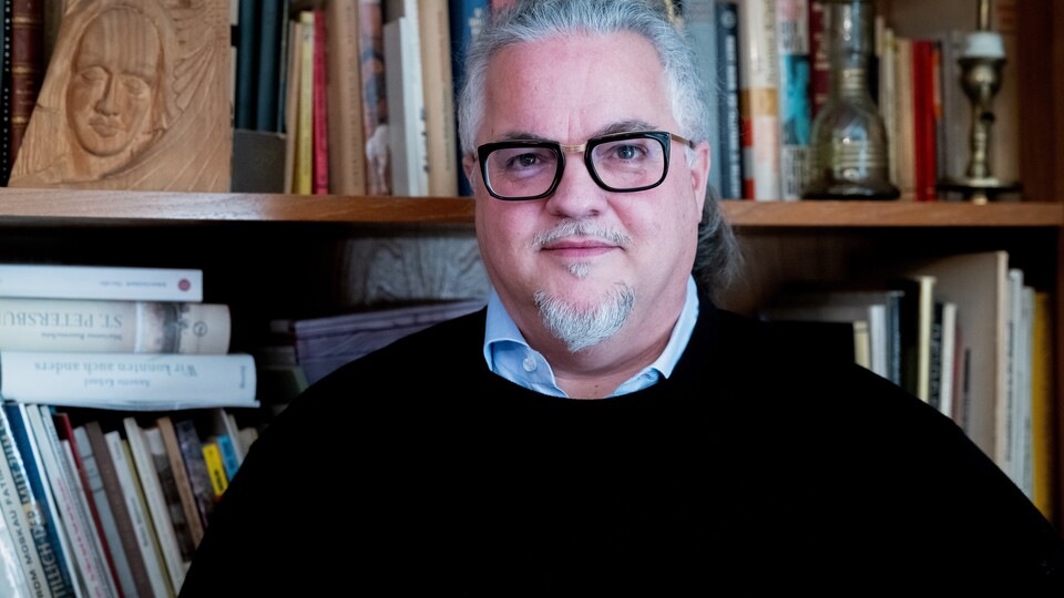 Porträt von Michael Meckel. Er trägt eine schwarze Brille und einen dunklen Pullover und schaut freundlich in die Kamera. Hinter ihm befindet sich ein Bücherregal.