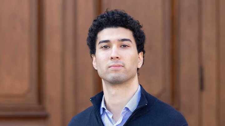 Mohamed Khalfallah. He is wearing a blue jacket over a blue shirt and is standing in front of brown door.
