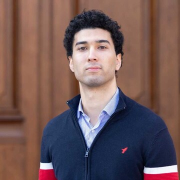 Mohamed Dhia Khalfallah. Er trägt eine blaue Jacke über einem weißen Hemd und steht vor einer Holztür.