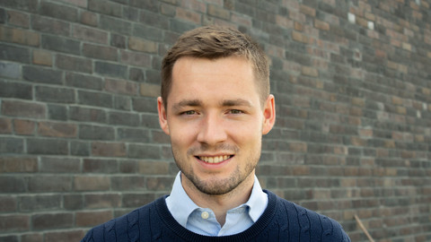 Nils Schweikard. He is wearing a blue sweater over a blue shirt and is standing in front of a grey brickstone wall.