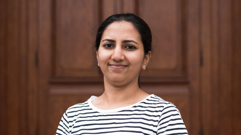 Samhitha Srinivas. She is wearing a black-white stripped sweater and is standing in front of a brown door.