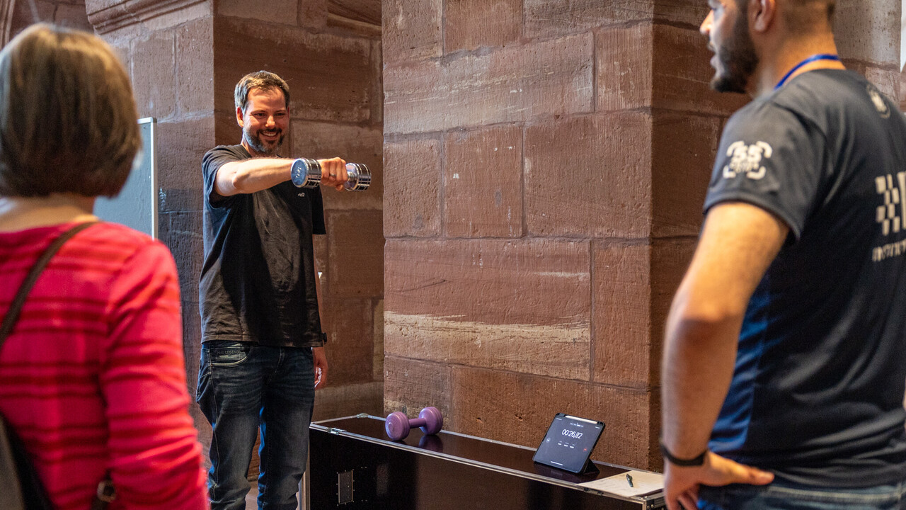 Ein Mann streckt seinen rechten Arm gerade nach vorne, in der Hand hält er eine Kurzhantel. Neben ihm steht eine schwarze Kiste, auf der sich eine weitere Kurzhantel und ein Tablet mit Stoppuhr-Anzeige befinden. Im Vordergrund stehen eine Frau und Mann mit dem Rücken zur Kamera und blicken in Richtung des Mannes mit der Hantel.