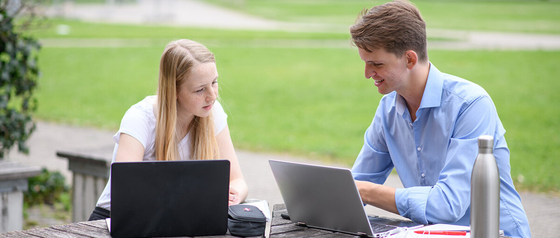 Ein Student und eine Studentin sitzen draußen an einem Tisch und lernen gemeinsam an ihren Laptops.