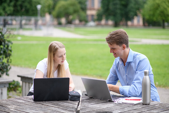 Ein Student und eine Studentin sitzen draußen an einem Tisch und lernen gemeinsam an ihren Laptops.