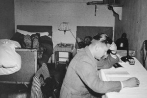 schwarz-weiß Aufahme eines Wohnbunkers am Goetheplatz. Ein kleiner Raum mit zwei Einzelbetten, auf denen Studierende liegen. Davor eine studierende Person, die an einem Schreibtisch sitzt.