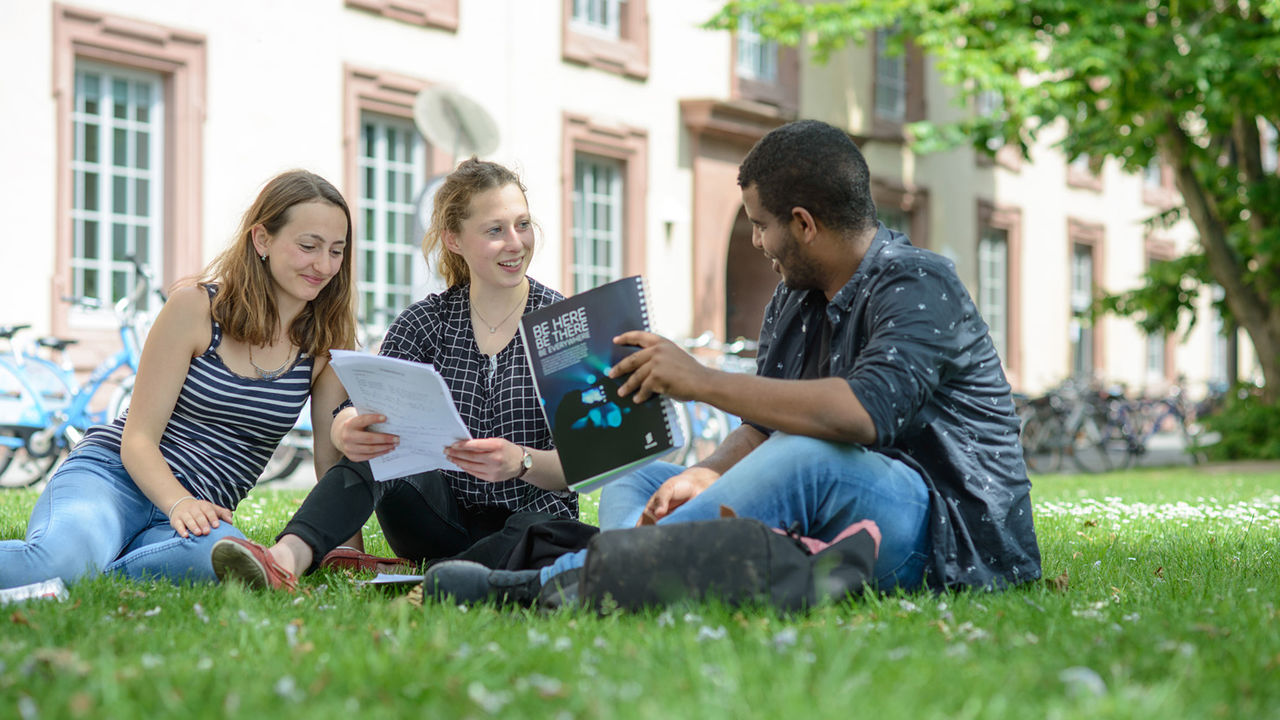 Zwei Studentinnen und ein Student sitzen auf einer Wiese vor dem Mannheimer Schloss. Sie tragen kurzärmlige Oberteile und halten Papierblöcke in ihren Händen.