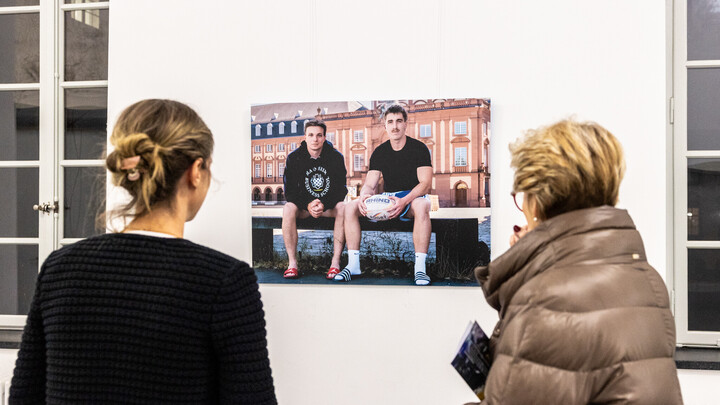Zwei Personen betrachten ein Foto an der Wand. Auf diesem sind zwei Spitzensportler zu sehen.