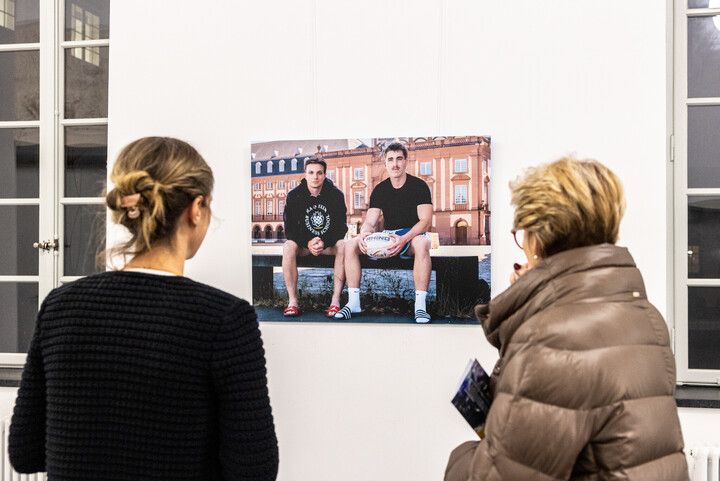 Zwei Personen betrachten ein Foto an der Wand. Auf diesem sind zwei Spitzensportler zu sehen.