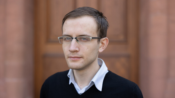 Andrii Tarasenko. He is wearing a black sweater over a white shirt and is standing in front of a brown door.