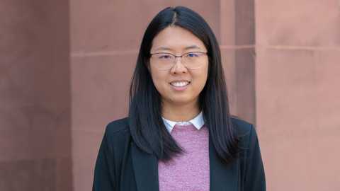 Yuhan Liu. She is wearing a black jacket over a white shirt and purple sweater and is standing in front of brown stone wall.