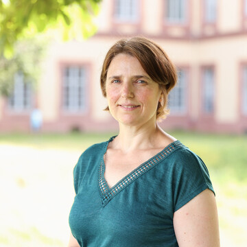 Katja Gutzmer hat braune kurze Haare. Sie trägt ein grünes T-Shirt mit V-Ausschnitt und ist auf einer grünen Wiese vor dem Schloss.  