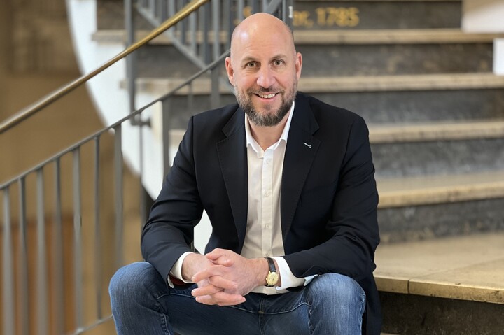 Patrick Barth sitzt auf einer Treppe im Schloss Mannheim. Er trägt ein weißes Hemd, ein schwarzes Sakko und eine Jeans. 