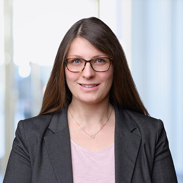 Katja Bauer hat lange braune Haare. Sie trägt eine braun umrandete Brille, einen dunkelgrauen Blazer und ein hellrosanes Oberteil.