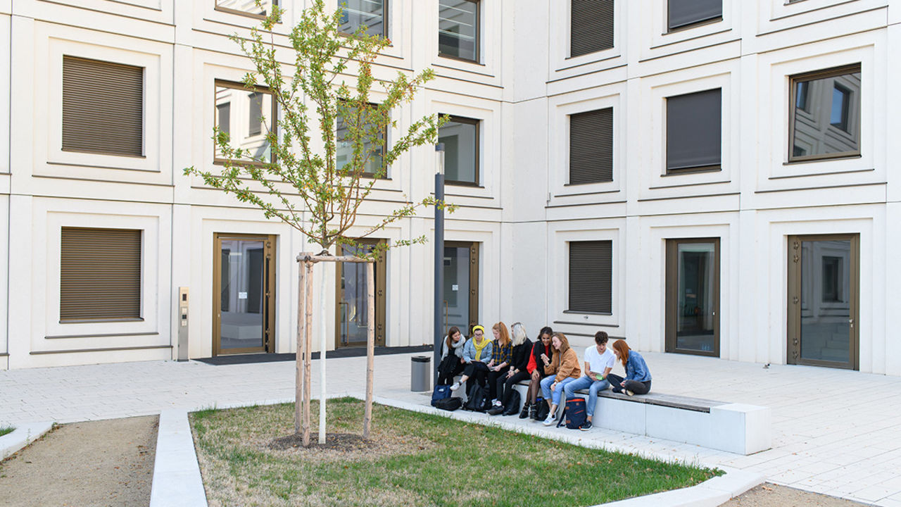 Studierende sitzen auf einer Bank und unterhalten sich. Im Hintergrund steht ein helles Gebäude mit vielen Fenstern.