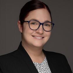 Porträt von Amelie Freiberg. Amelie Freiberg trägt einen schwarzen Blazer und eine weiße Bluse mit schwarzen Punkten. Die dunklen Haare hat sie zu einem Zopf zusammengebunden. Sie trägt eine Brille und lächelt selbstbewusst in die Kamera. 