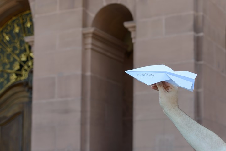 Eine Hand hält einen Papierflieger. Auf dem Papierflieger steht in die Luft. Auf dem Papierflieger ist ein Pfeil in die mögliche Flugrichtung abgebildet sowie die Aufschrift "VWL-Studium".