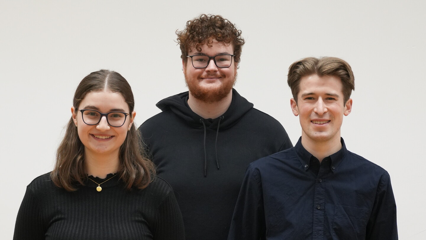 Drei schwarzgekleidete Personen stehen vor einer weißen Wand.