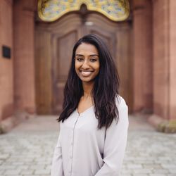 Rahel Habteab hat schwarze mittellange Haare und lächelt. Sie trägt eine weiße Bluse und steht vor dem Schloss auf dem Ehrenhof.