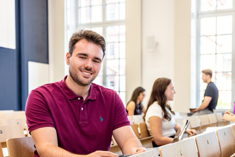 Ein Student sitzt in einer Bankreihe im Hörsaal. Er schaut direkt in die Kamera und lächelt. Im Hintergrund sitzen weitere Studierende.