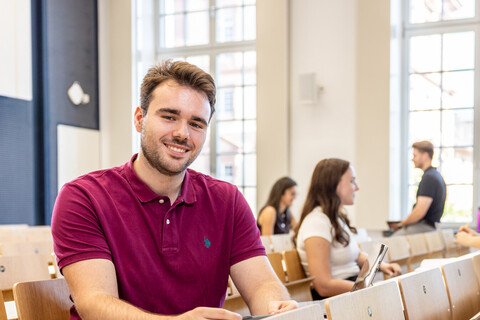 Ein Student sitzt in einer Bankreihe im Hörsaal. Er schaut direkt in die Kamera und lächelt. Im Hintergrund sitzen weitere Studierende.
