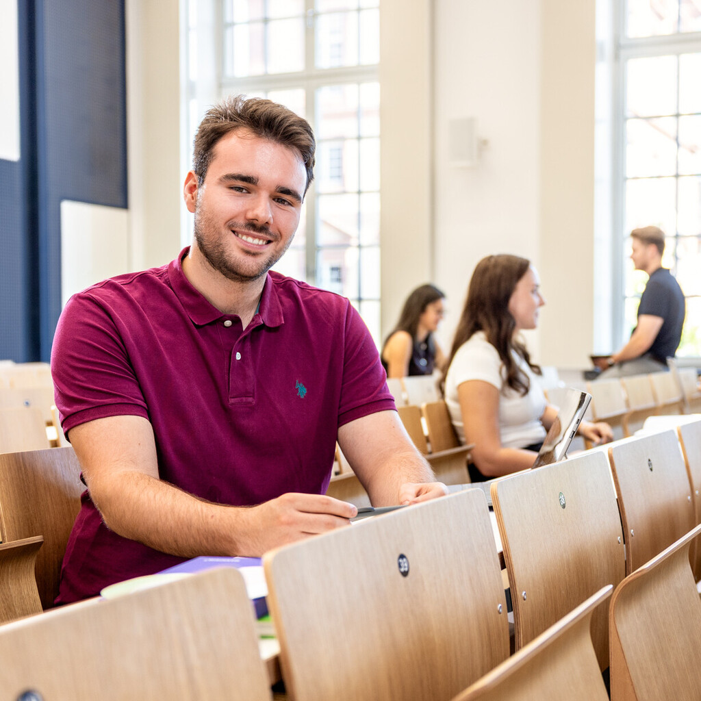 Ein Student sitzt in einer Bankreihe im Hörsaal. Er schaut direkt in die Kamera und lächelt. Im Hintergrund sitzen weitere Studierende.