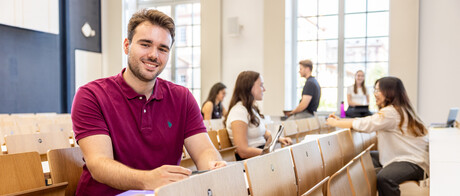 Ein Student sitzt in einer Bankreihe im Hörsaal. Er schaut direkt in die Kamera und lächelt. Im Hintergrund sitzen weitere Studierende.