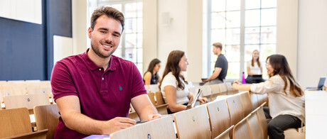 Ein Student sitzt in einer Bankreihe im Hörsaal. Er schaut direkt in die Kamera und lächelt. Im Hintergrund sitzen weitere Studierende.
