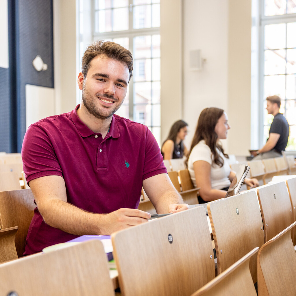 Ein Student sitzt in einer Bankreihe im Hörsaal. Er schaut direkt in die Kamera und lächelt. Im Hintergrund sitzen weitere Studierende.