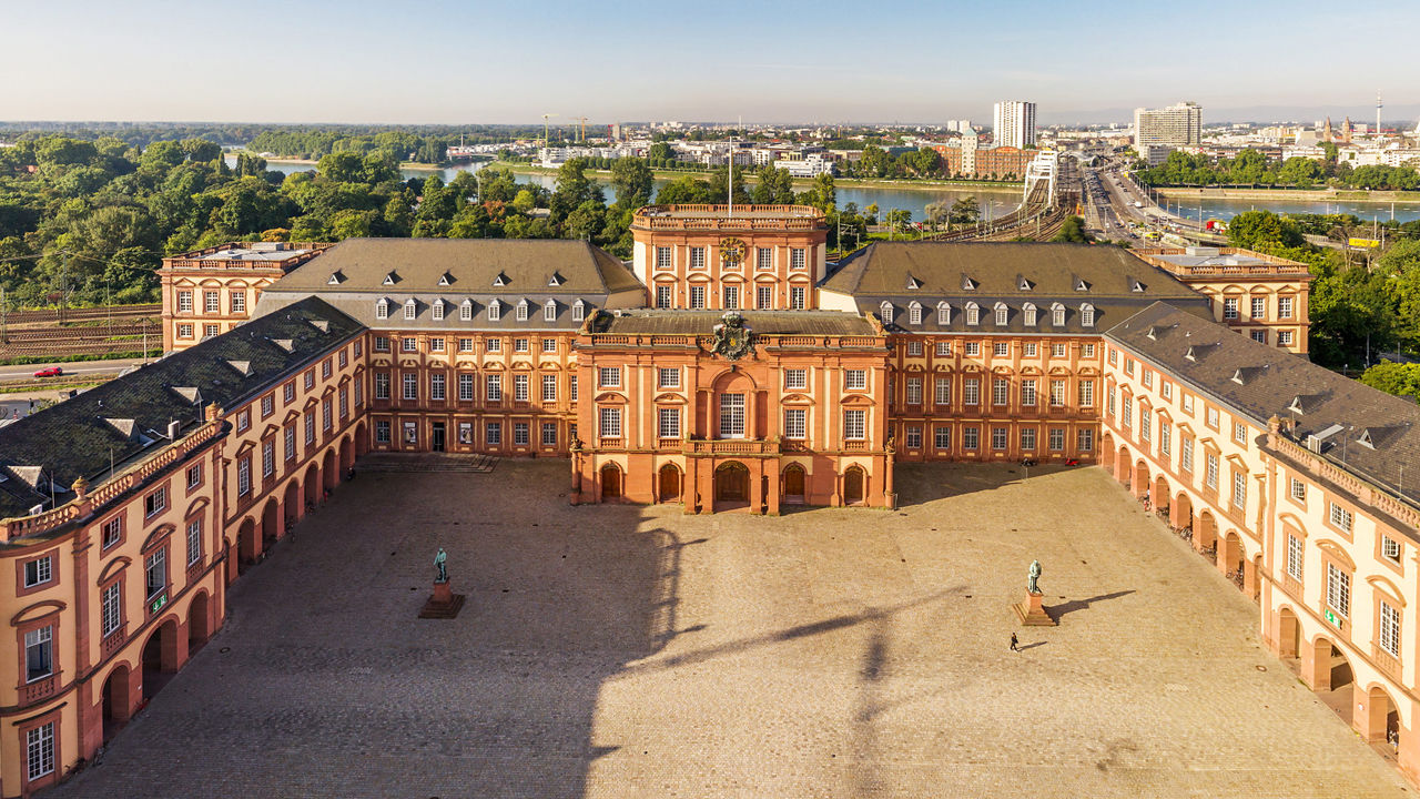 Das Mannheimer Schloss von oben. Die helle Fassade mit den vielen Fenstern leuchtet im Sonnenlicht. Im Hintergrund fließt der Rhein.