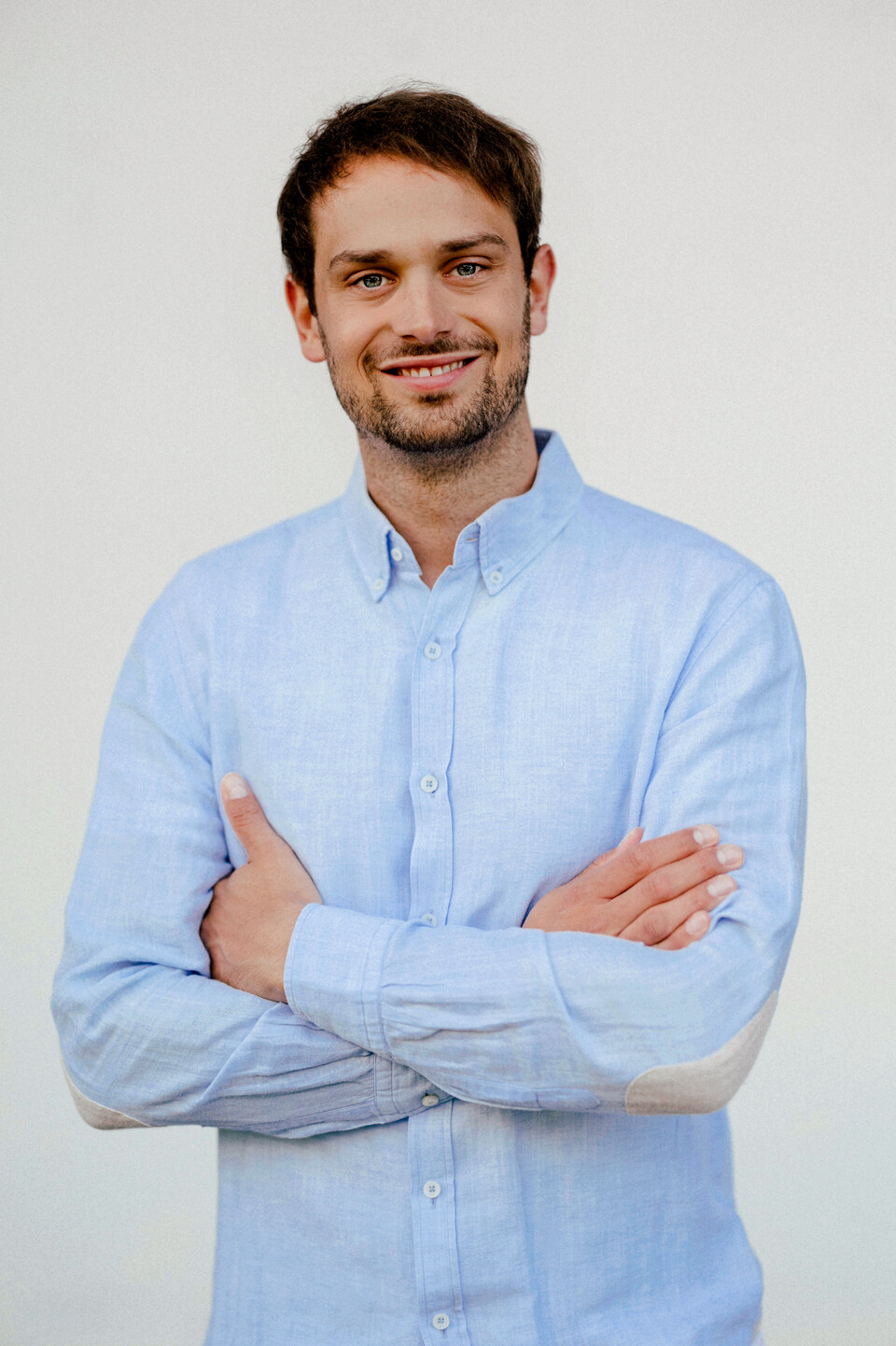 Martin Glanzer trägt ein hellblaues. langärmeliges Hemd und lächelt. Seine Arme sind verschränkt.
