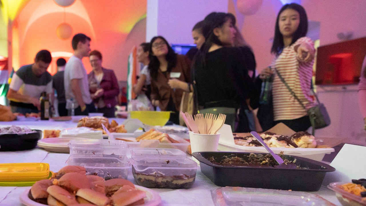 Auf einem Tisch stehen viele verschiedene internationale Speisen. Viele Menschen schauen vergnügt auf das Essen.