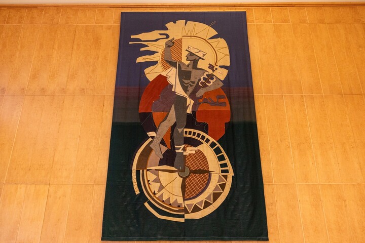 Darstellung des Götterboten Hermes auf einem großformatigen Teppich, mit Heroldstab und geflügelten Sandalen. Im Hintergrund drei runde Formen: Oben eine angedeutete Sonne, unten ein Kompass.