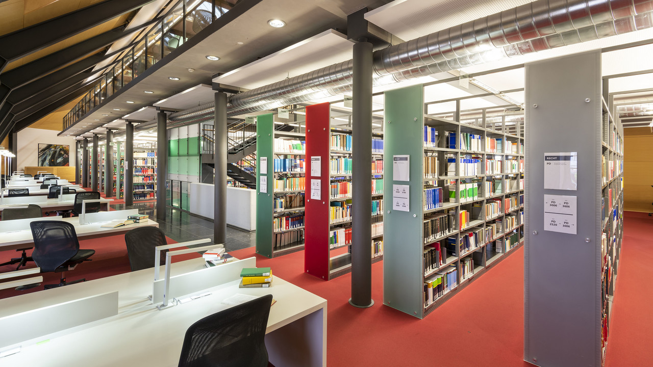In der Ehrenhof-Bibliothek ist ein roter Teppichboden. Viele große Bücherregale stehen in der Mitte des Raumes, an der Seite sind Schreibtische.