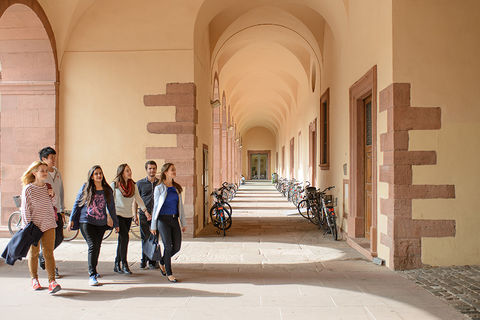 Vier Studentinnen und zwei Studenten spazieren über den Campus der Universität Mannheim. Die Studierenden sind frühlingshaft gekleidet. In einem Gang im Hintergrund stehen viele Fahrräder an den sandfarbenen Wänden des Schlosses.